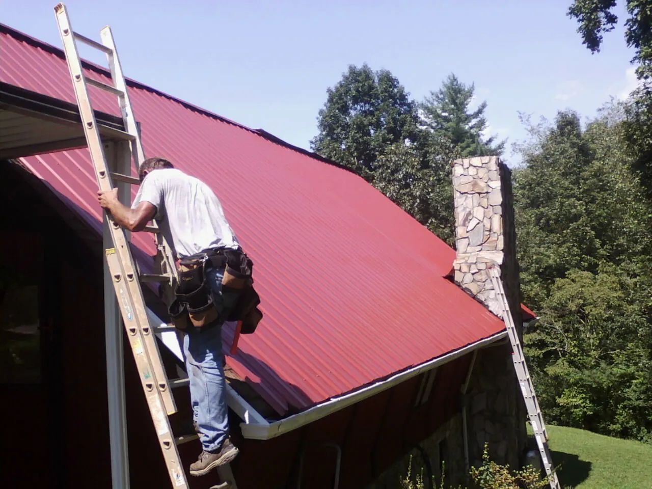 Roofer on a ladder beside a red metal roof and stone chimney — Soffit & Fascia Repair by Roofing Brothers in Feeding Hills